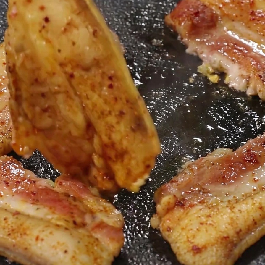 Pinzas de metal volteando un trozo de panceta de cerdo dorada en una sartén parrilla caliente.