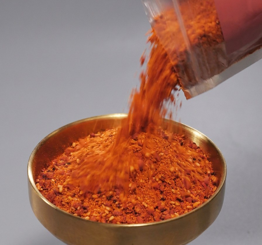 Condimento en polvo rojo con semillas de sésamo vertido desde un paquete de plástico en un cuenco de color bronce.
