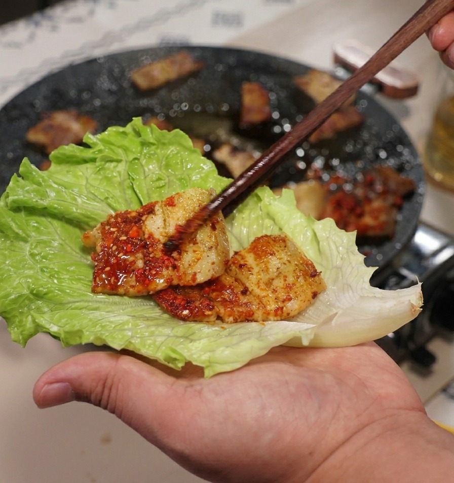 Una mano sosteniendo una hoja grande de lechuga verde mientras coloca un trozo de panceta de cerdo con palillos.