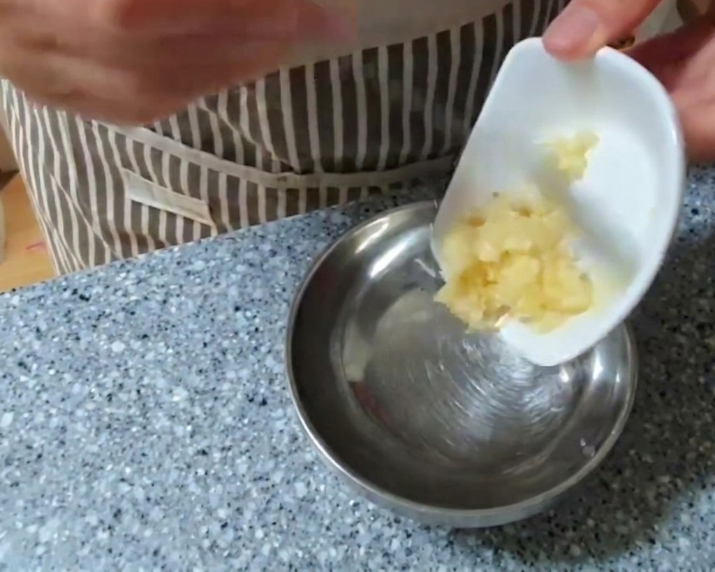 Una persona añadiendo ajo picado desde un pequeño plato blanco a un bol grande de acero inoxidable.