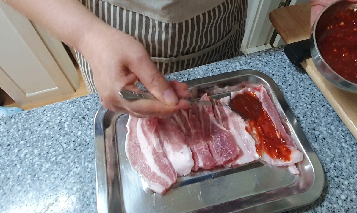 Una mano usando una cuchara para cubrir tiras de panceta cruda con un marinado espeso de chile rojo brillante sobre una bandeja plateada.