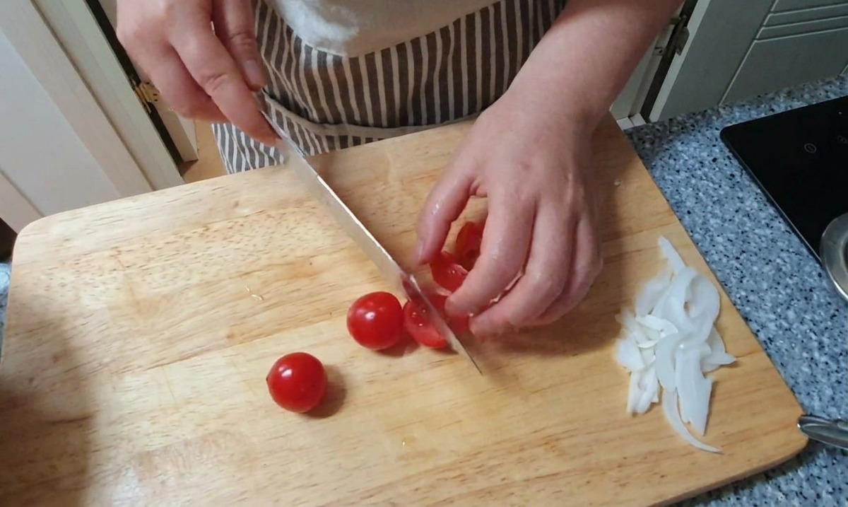 Cortando pequeños tomates cherry rojos por la mitad sobre una tabla de madera junto a un montón de cebolla blanca picada.