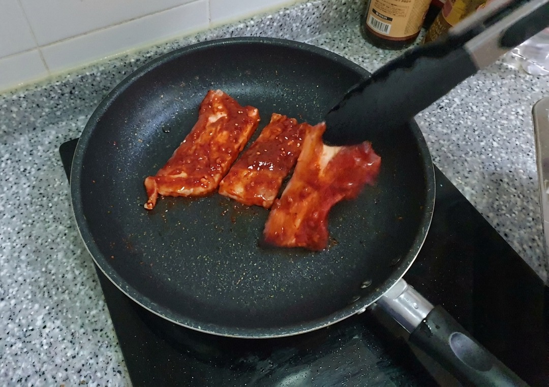 Tiras de panceta marinadas de color rojo colocándose en una sartén antiadherente negra con la ayuda de unas pinzas.