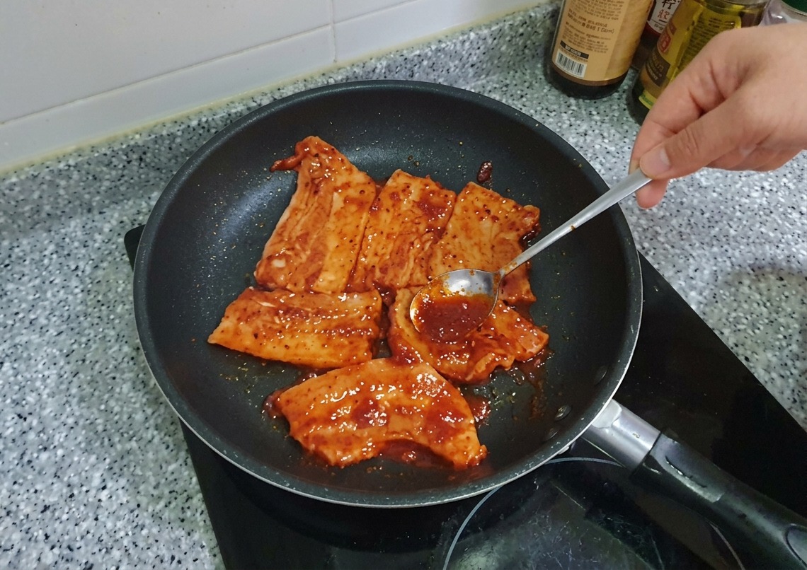 Una cuchara extendiendo un espeso marinado rojo de Gochujang sobre rodajas de panceta de cerdo en una sartén.