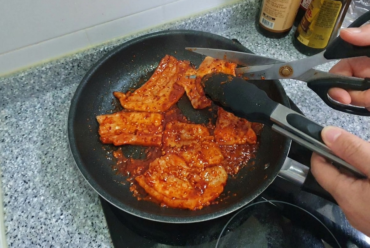 Tijeras de cocina cortando tiras de panceta de cerdo picante cocinada dentro de una sartén.
