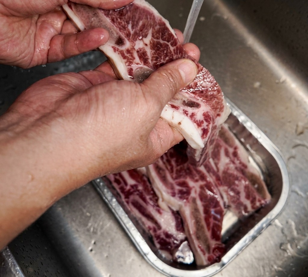 Una mano sosteniendo un trozo de costilla de res marmoleada bajo un chorro de agua fría en un fregadero de acero inoxidable.