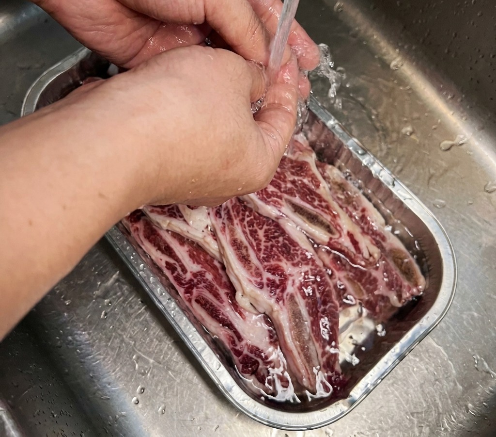 Costillas de res sumergidas en una bandeja de metal que se llena con agua fría limpia desde un grifo.
