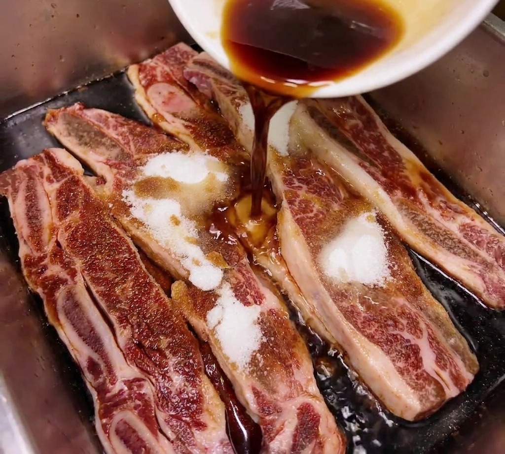 Salsa de soja oscura o mirin cayendo desde un tazón blanco sobre costillas de res crudas espolvoreadas con azúcar en una bandeja metálica.