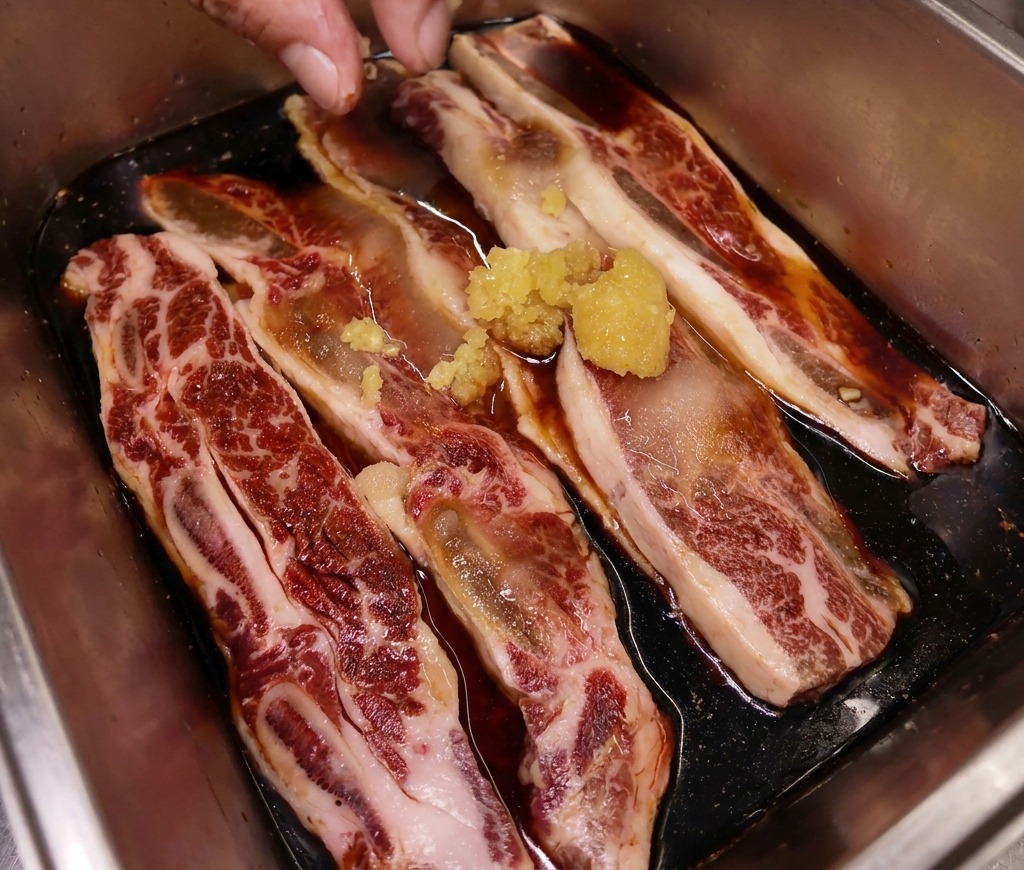 Una mano colocando un pequeño montón de jengibre fresco picado sobre costillas de res que se marinan en una bandeja de acero inoxidable.