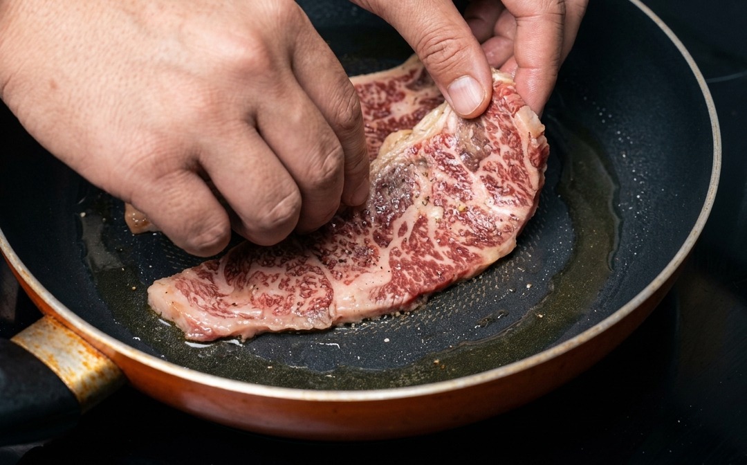 Una mano colocando una costilla de res marmoleada y marinada en una sartén caliente.