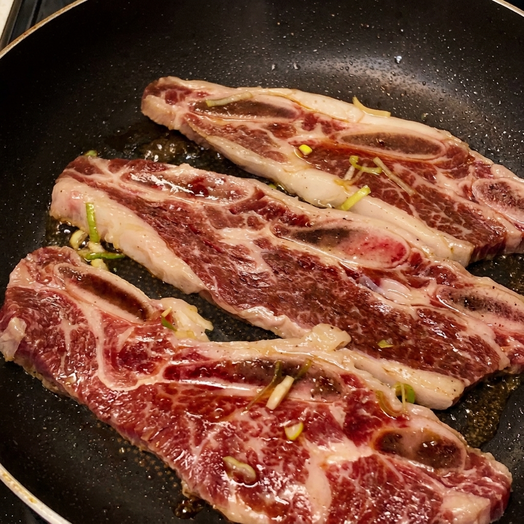 Tres costillas de res chisporroteando en una sartén mientras el marinado oscuro burbujea y se reduce en un glaseado espeso y brillante.