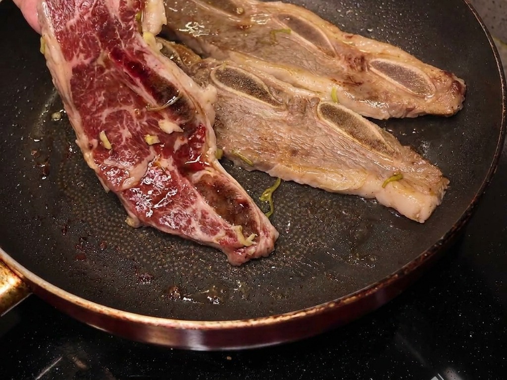Una mano volteando una costilla de res a medio cocinar en una sartén, mostrando una superficie caramelizada y dorada.