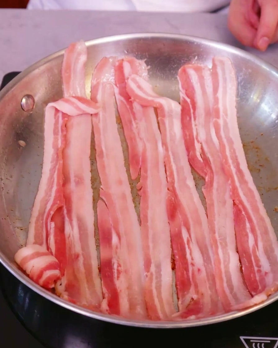 Lonchas de panceta de cerdo fresca dispuestas en una sartén grande de acero inoxidable sobre una placa de inducción.