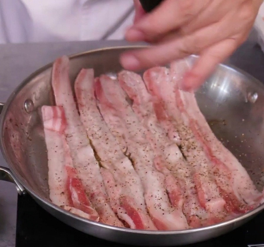 Sazonando lonchas de panceta cruda con un molinillo de pimienta negra en una sartén de acero inoxidable.