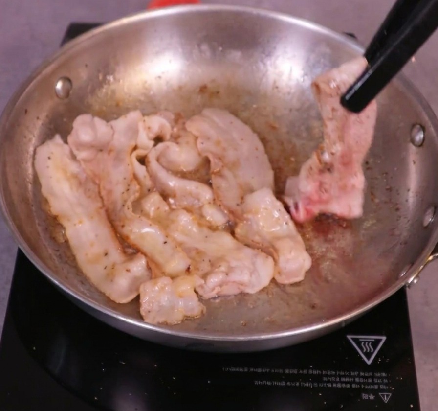 Lonchas de panceta doradas dadas la vuelta en una sartén caliente con la ayuda de unas pinzas de cocina negras.