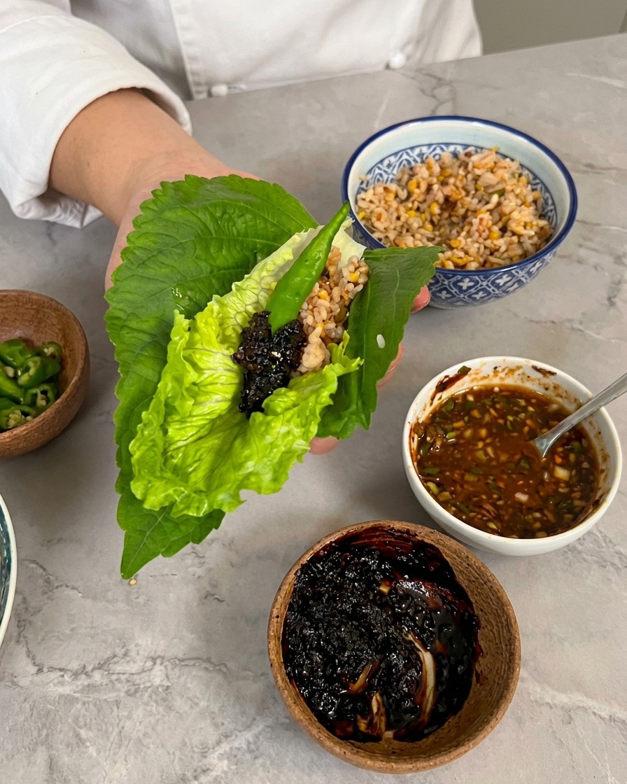 Una mano sosteniendo un wrap de lechuga y hoja de perilla cubierto con una cucharada de salsa oscura y un chile verde.