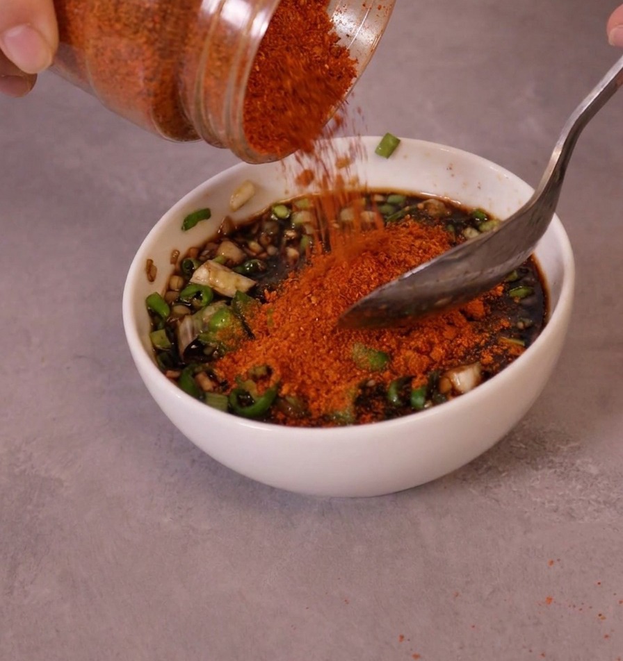 Chile en polvo fino de color rojo brillante vertiéndose desde un tarro a un cuenco con la base de salsa y chiles.