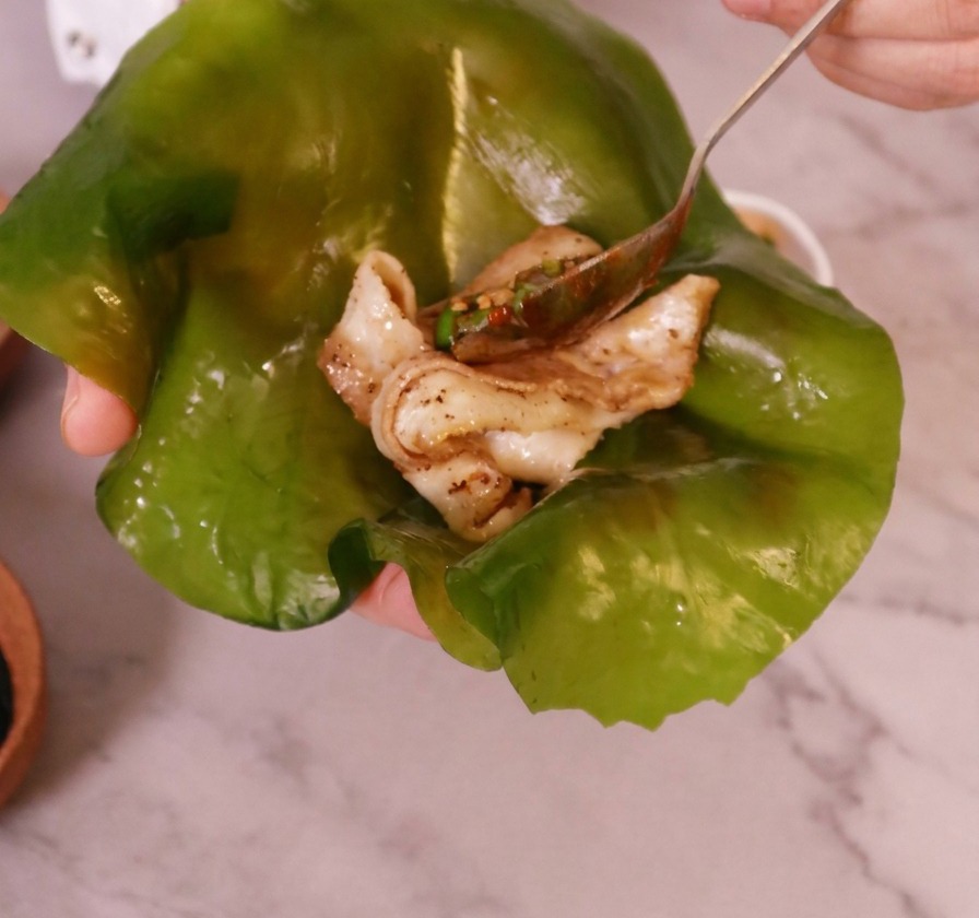 Una mano sosteniendo una lámina grande y translúcida de alga kelp verde con un trozo de panceta cocinada en el centro.
