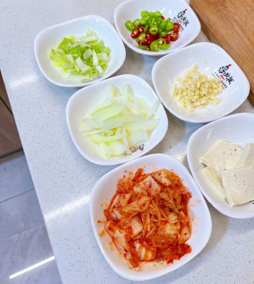 Varios cuencos blancos pequeños con cebolletas picadas, pimientos verdes y rojos, ajo picado, cebolla rebanada, tofu y kimchi.