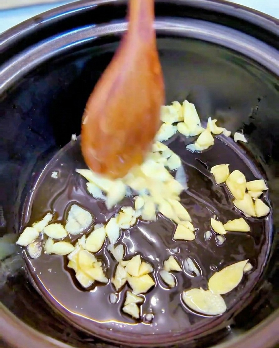 Ajo picado mezclándose con aceite de sésamo en el fondo de una olla negra con una cuchara de madera.