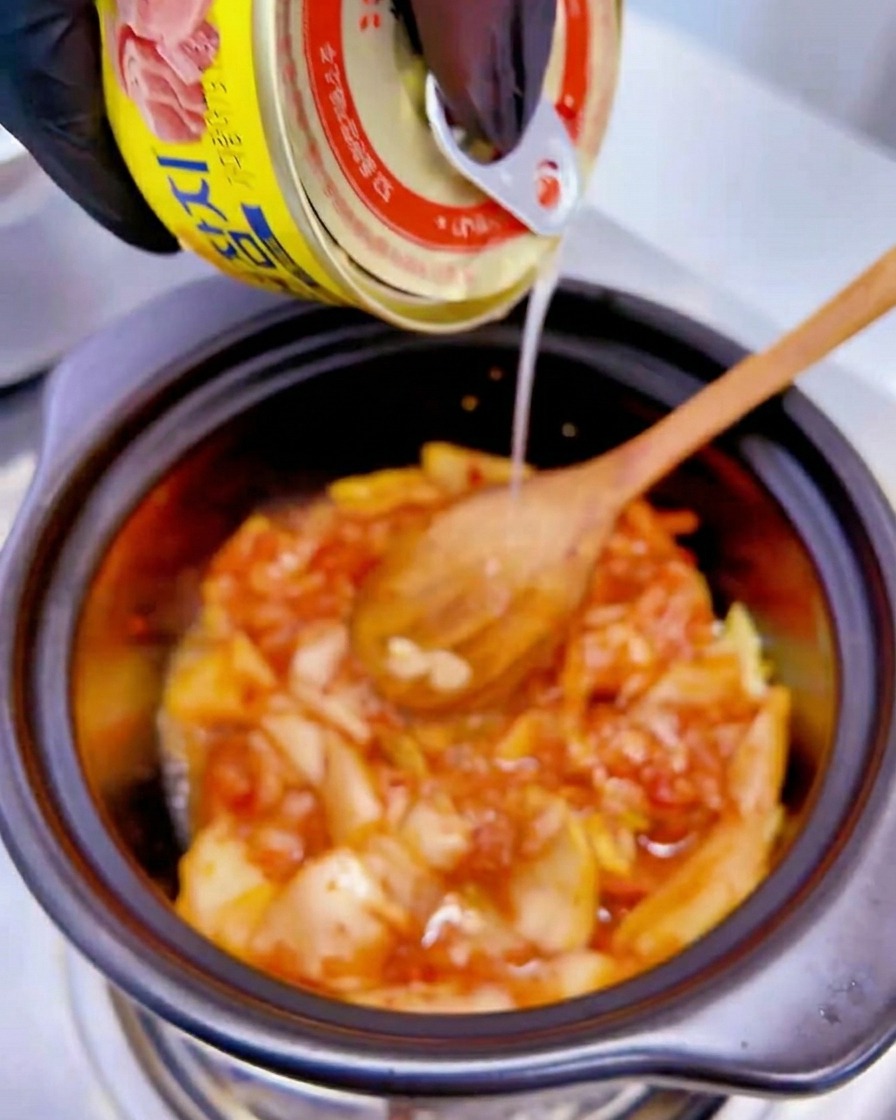 Líquido de una lata amarilla de atún vertiéndose sobre la mezcla de kimchi en la olla.