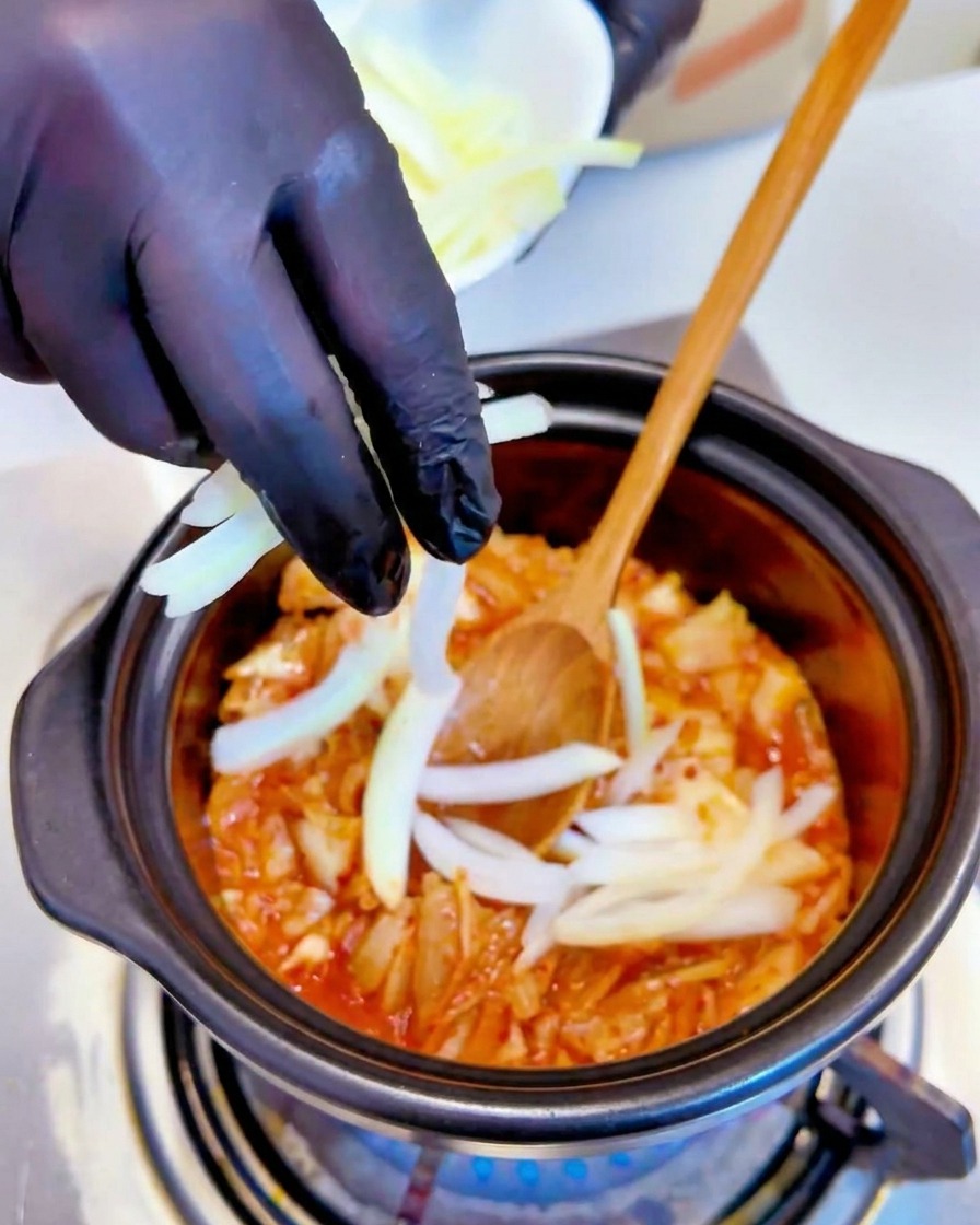 Cebolla blanca rebanada agregándose a la mezcla de kimchi hirviendo en una olla por una mano con guante negro.