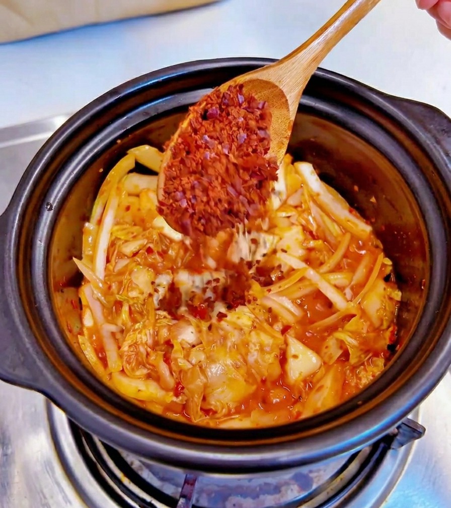 Una cuchara de madera agregando copos de chile rojo grueso a una olla negra con kimchi salteado y cebolla rebanada.