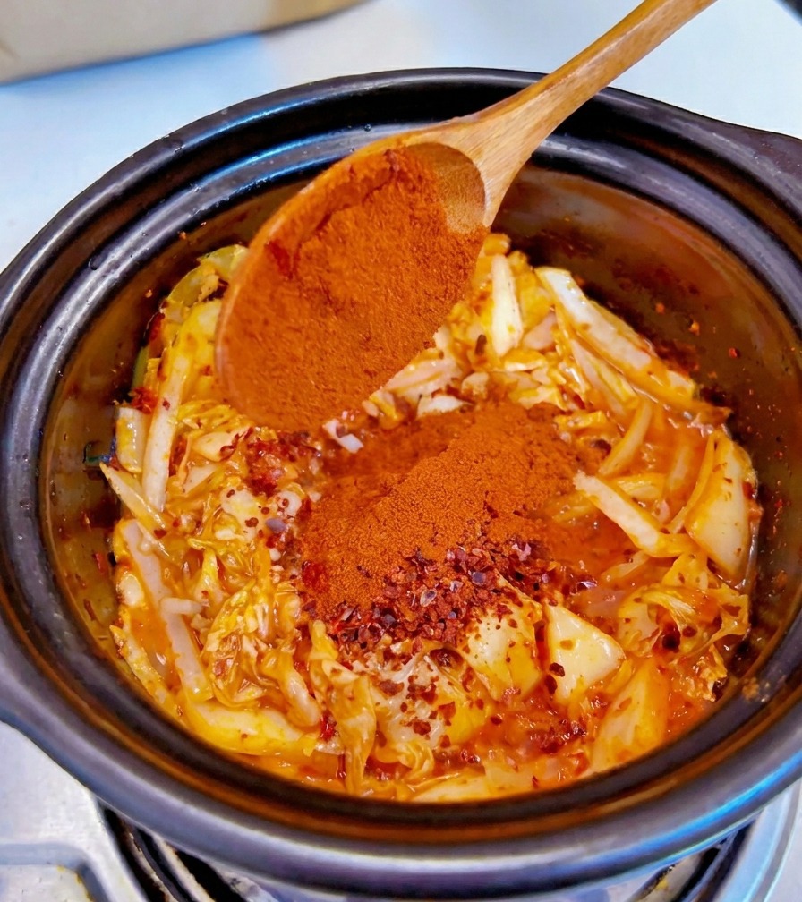 Chile en polvo rojo fino cayendo desde una cuchara de madera sobre la mezcla sazonada de kimchi y cebolla.