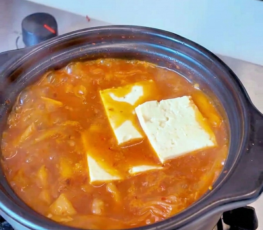 Rebanadas rectangulares de tofu blanco sumergidas en un caldo rojo de estofado de kimchi hirviendo.