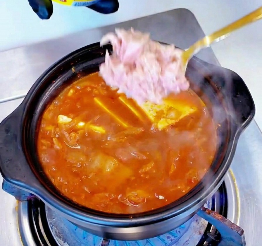 Trozos de atún claro en lata colocándose con una cuchara en el centro de un estofado rojo que hierve a fuego lento.