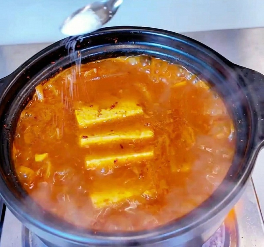 Una pequeña cuchara de metal espolvoreando sal blanca sobre la superficie de un estofado de kimchi y atún burbujeante.
