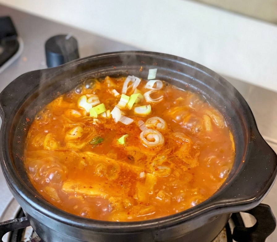 Un estofado de kimchi y atún rojo y burbujeante en una olla tradicional de barro negro sobre la estufa, decorado con cebolletas frescas y rodajas de pimiento.