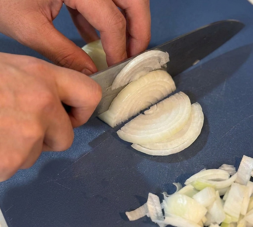 Manos usando un cuchillo de chef para cortar finamente una cebolla blanca en una tabla de picar azul.