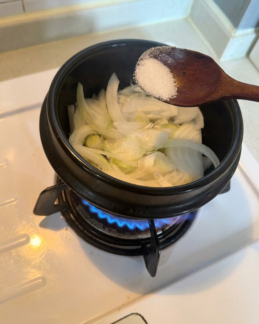 Cebolla blanca en rodajas dentro de una olla, sazonada con una cucharada de azúcar blanca sobre un quemador de gas encendido.