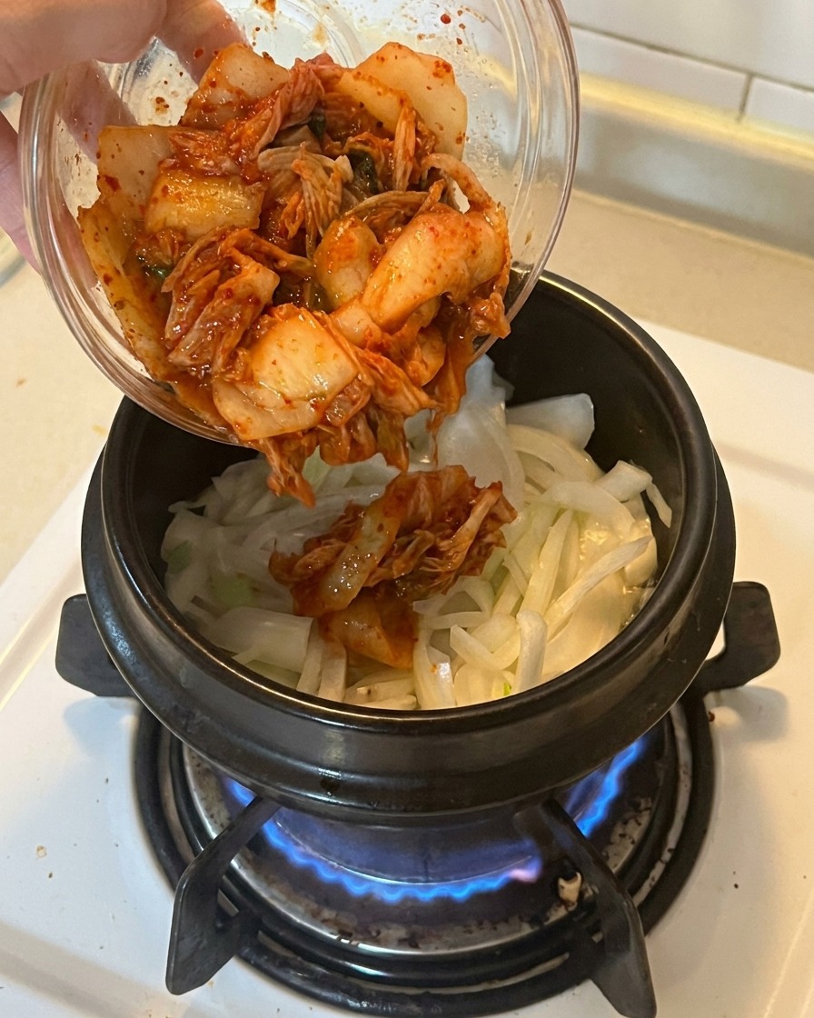Añadiendo un tazón de kimchi picado de color rojo vibrante en una olla que contiene cebollas y cebolletas sofritas.