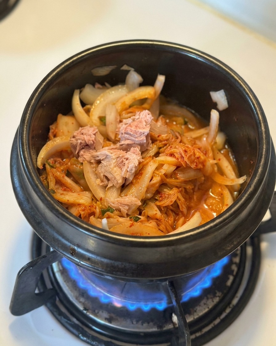 Trozos de atún en lata colocados sobre una cama de kimchi y cebollas sofritas en una olla negra.