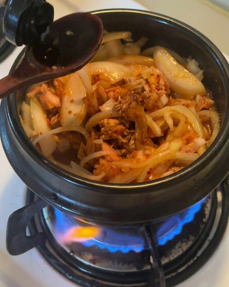 Una cuchara de madera vertiendo salsa de soja oscura en una olla de barro negra que contiene cebollas, atún y kimchi salteados.