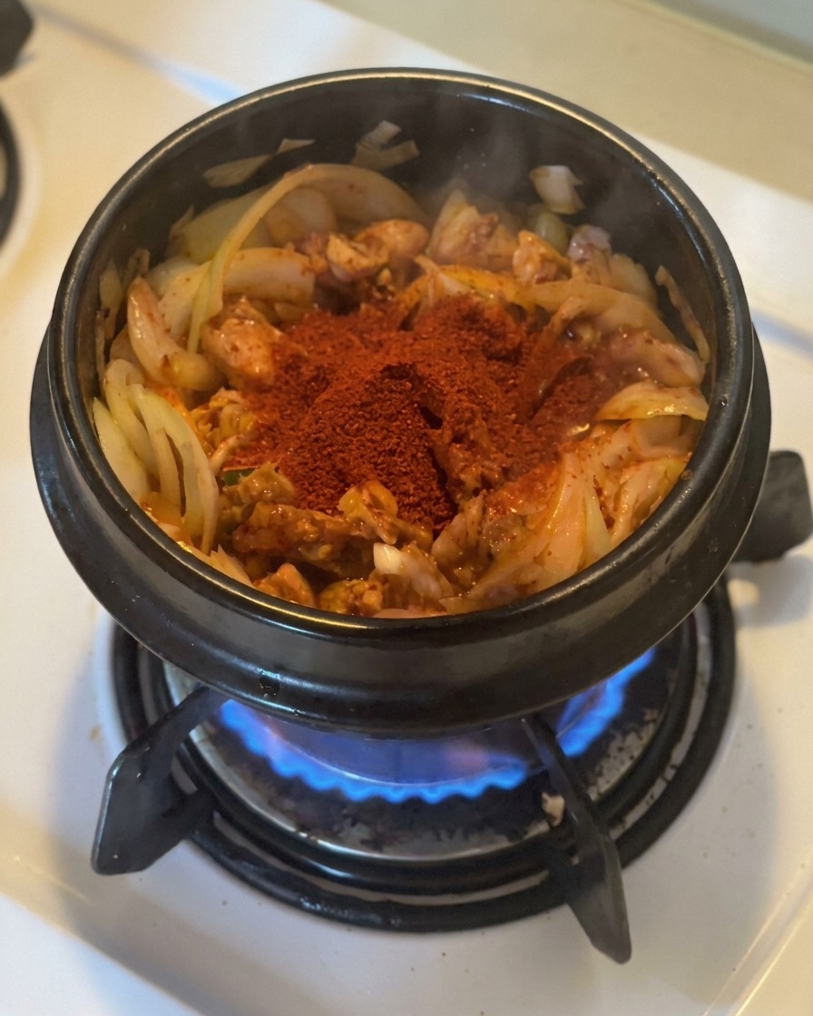 Una porción generosa de gochugaru rojo brillante sobre el kimchi y las cebollas salteadas en una olla de piedra negra sobre una llama de gas.