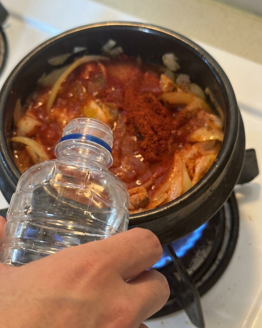Agua vertiéndose desde una botella de plástico transparente en una olla llena de kimchi sazonado de rojo y atún.