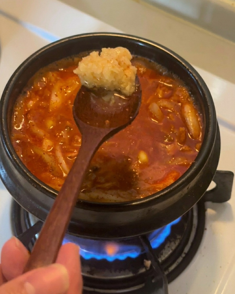 Una cuchara de madera sosteniendo una porción de ajo picado sobre un estofado de kimchi y atún de color rojo burbujeante.