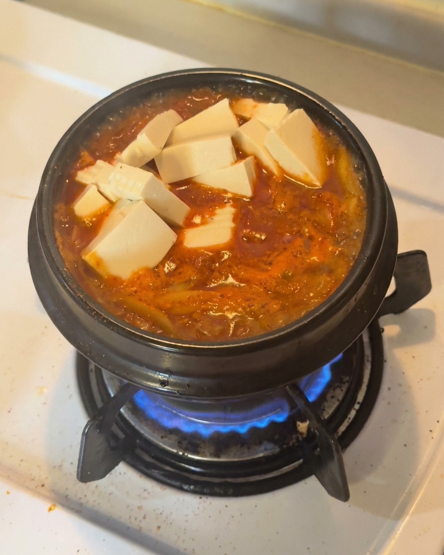 Cubos de tofu blanco fresco y rodajas de chile verde acomodados sobre un estofado de kimchi rojo que burbujea en una olla de barro negra.