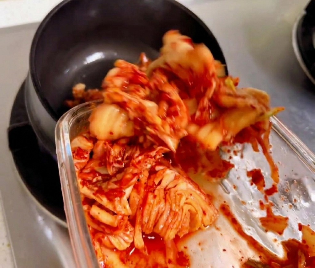 Kimchi picado siendo salteado y removido en una cazuela tradicional de piedra negra sobre una llama de gas.