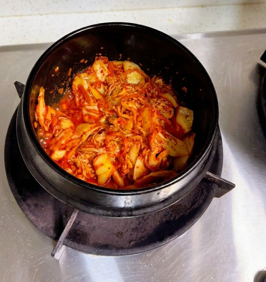 Una mano vertiendo agua de arroz turbia en una cazuela de piedra que contiene el kimchi salteado.