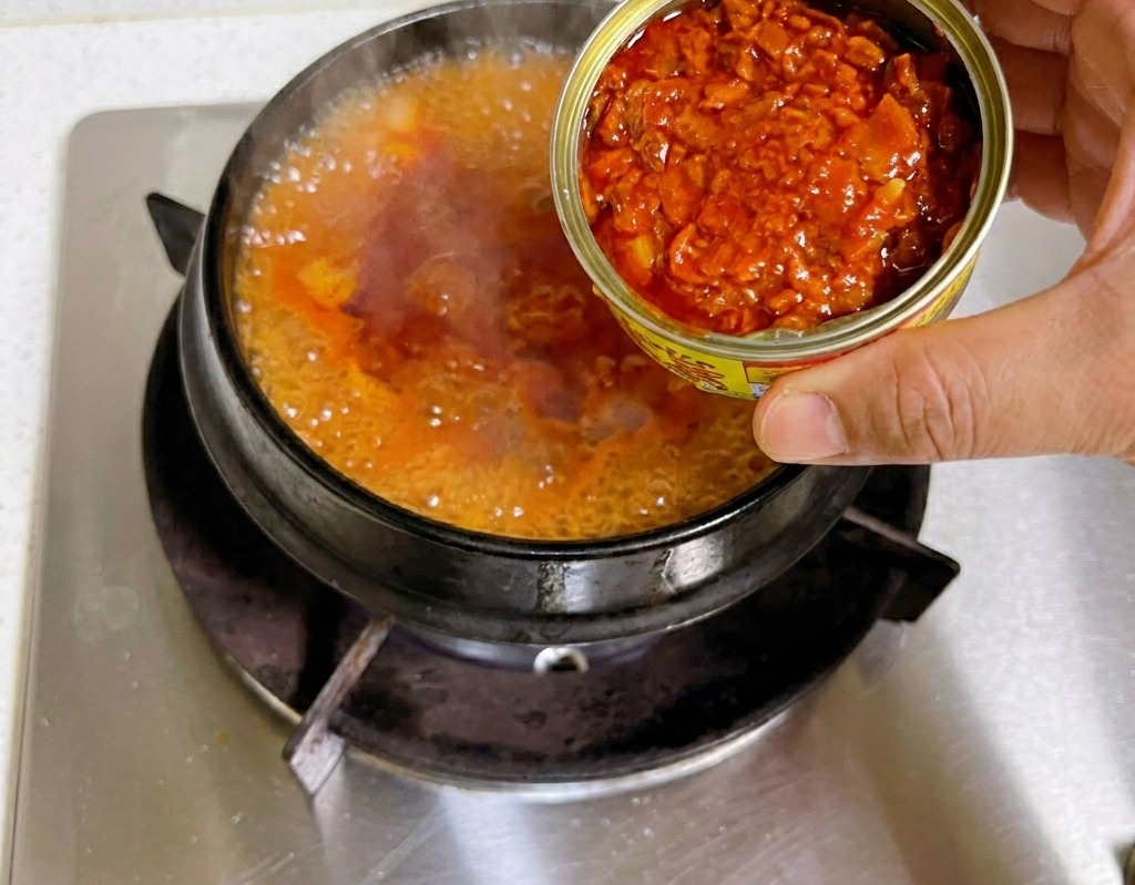 Una mano sosteniendo una lata de conserva abierta y vertiendo trozos de atún en una base de estofado de kimchi rojo que hierve en una cazuela de piedra.