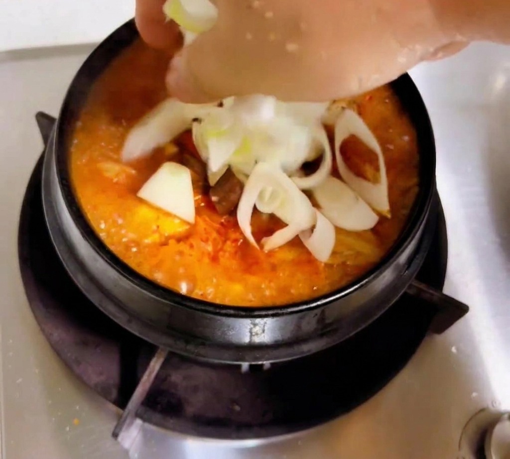 Una mano agregando rodajas de cebolla blanca y cebollines a un estofado de kimchi de color naranja rojizo que hierve a fuego lento.