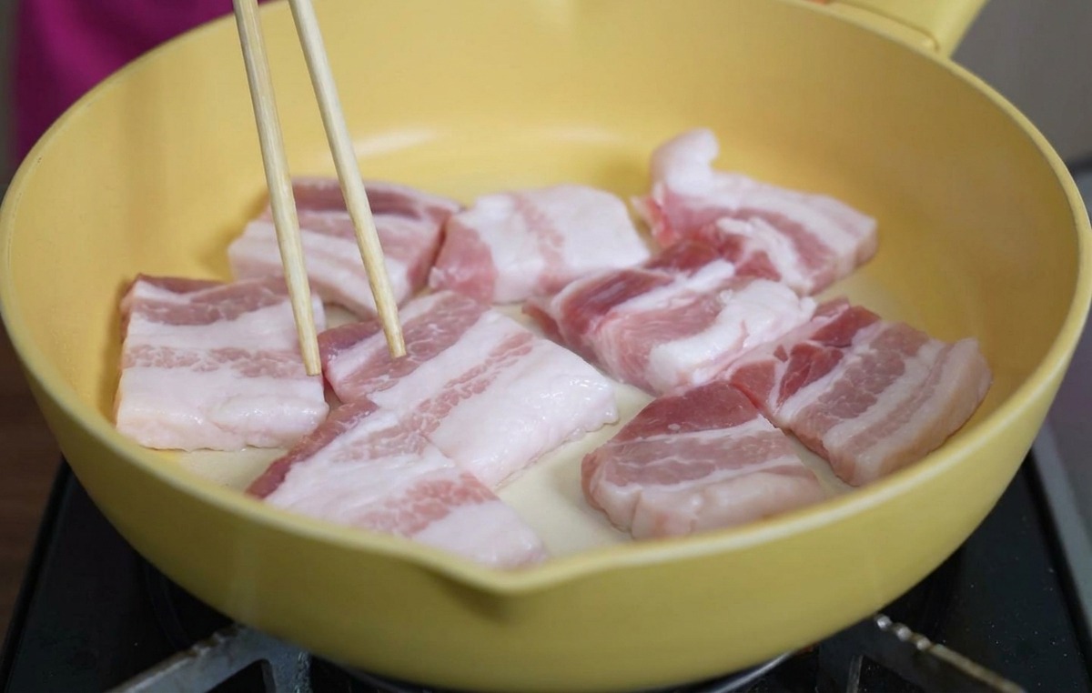 Lonchas gruesas y crudas de panceta dispuestas en una sartén de cerámica amarilla mientras se acomodan con palillos.