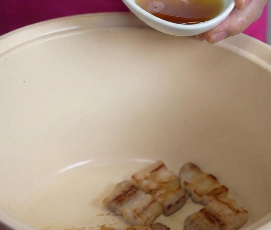 Un pequeño cuenco blanco vertiendo aceite de sésamo dorado sobre los trozos de panceta frita en una gran olla de color crema.