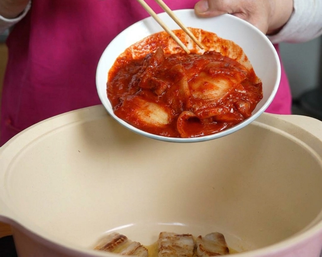 Una mano usando palillos para deslizar un tazón de kimchi rojo picante en una olla que contiene la panceta de cerdo.