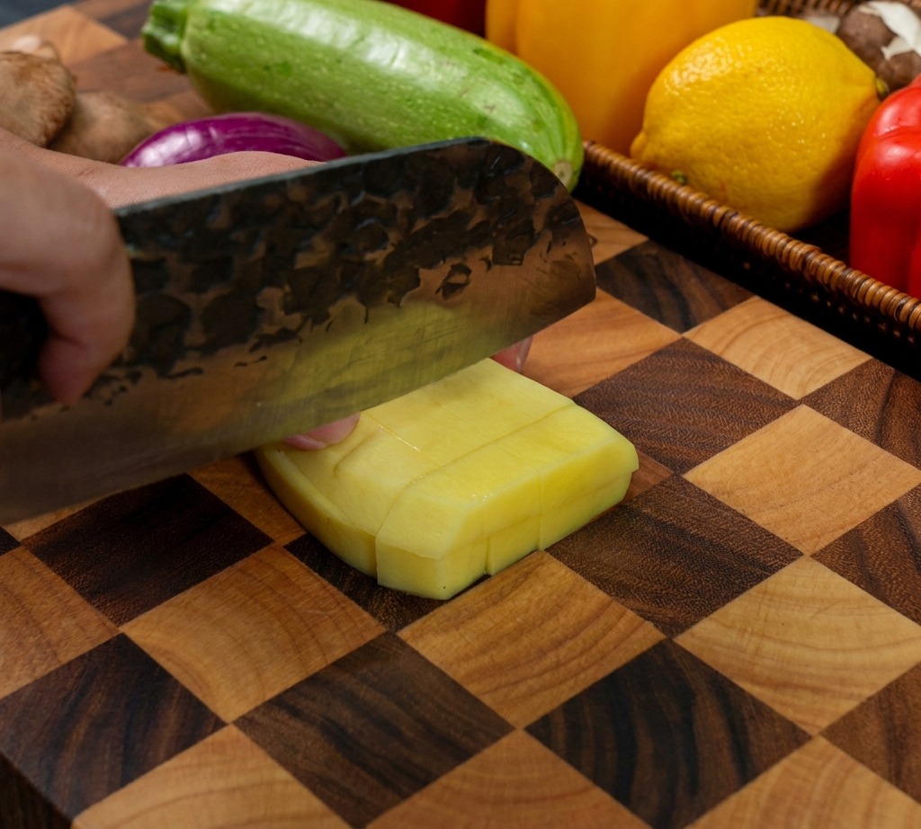 Un cuchillo afilado cortando una patata amarilla pelada para crear cubos pequeños y uniformes sobre una tabla de cortar de madera.