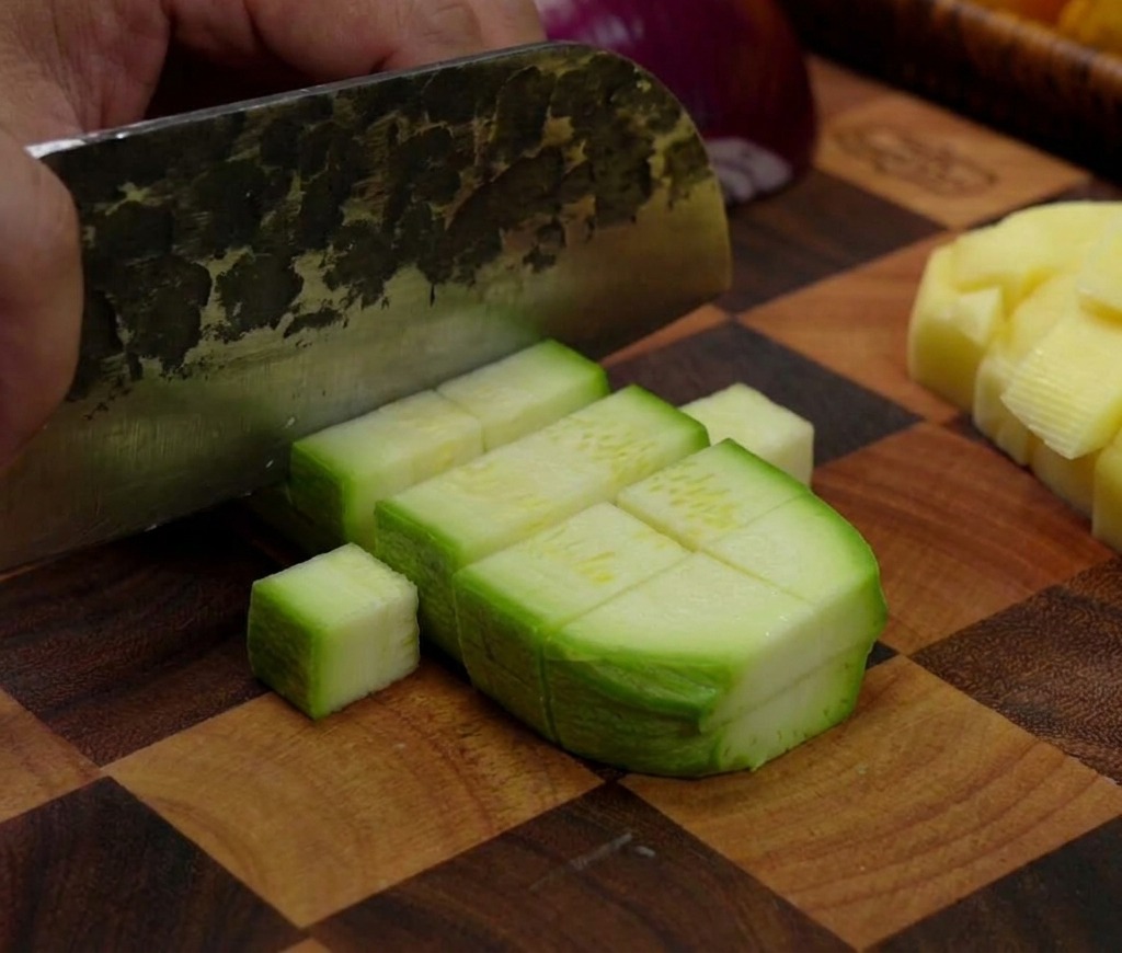 Un hacha de cocina cortando un calabacín verde, dividiéndolo en pequeños trozos rectangulares sobre una tabla de madera a cuadros.